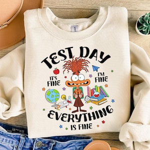 May include: A beige sweatshirt with a cartoon character wearing a backpack and holding books. The text on the sweatshirt reads "Test Day, It's Fine, I'm Fine, Everything is Fine."
