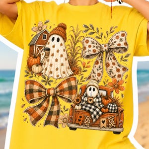May include: A bright yellow t-shirt featuring a graphic print of two ghosts, one wearing an orange beanie and holding a coffee cup, the other sitting in the back of a truck. The design includes pumpkins, a barn, floral bows, and plaid patterns.