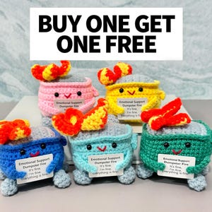 May include: A collection of colorful, crocheted "Emotional Support Dumpster Fire" toys. Each toy is a different color, including pink, yellow, blue, teal, and green, with a smiling face and a flame on top. Each toy holds a small sign that reads "It's fine, I'm fine, everything is fine."