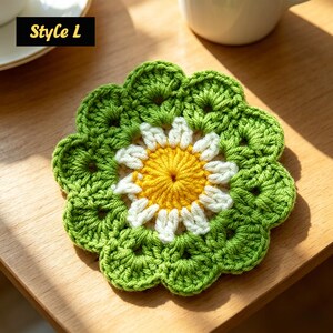 May include: A crocheted flower-shaped coaster with a yellow centre, white petals, and green leaves. The text "Style L" is in a black box in the upper left corner. The coaster is on a wooden surface.