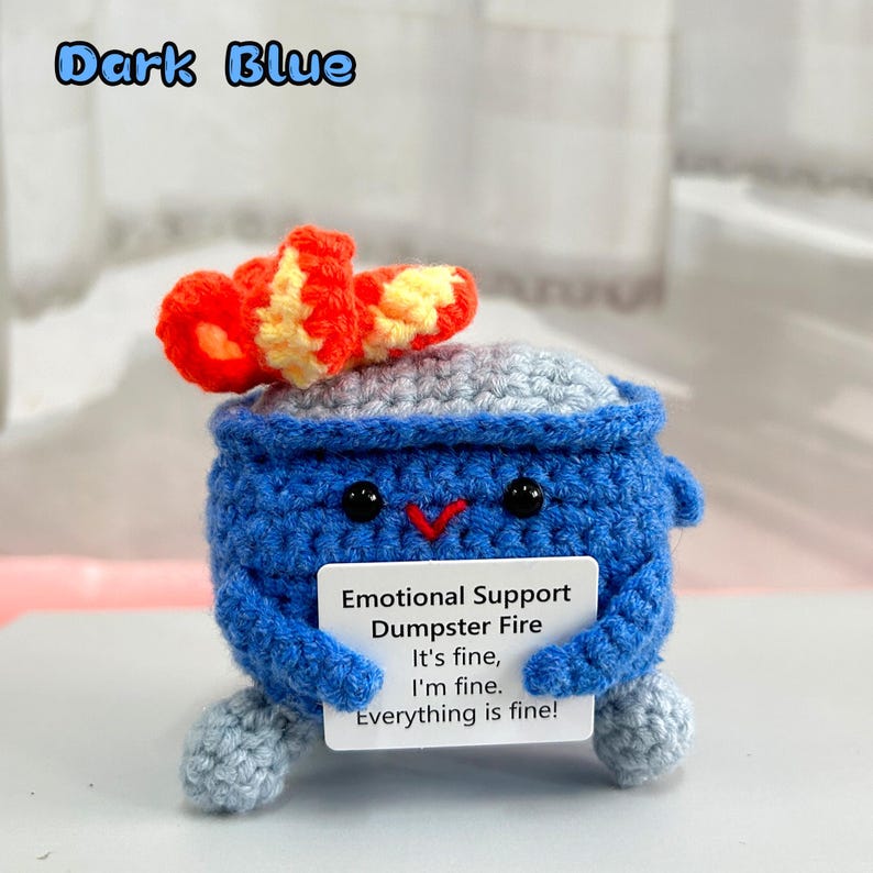 May include: A crocheted blue dumpster fire with a red and yellow flame on top. The dumpster fire is holding a sign that says "Emotional Support Dumpster Fire It's fine, I'm fine. Everything is fine!"