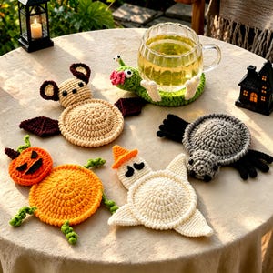 Könnte beinhalten: Ein Set aus fünf gehäkelten Untersetzern mit Halloween-Motiven, darunter eine beige Fledermaus, eine grüne Schnecke, ein orangefarbener Kürbis, ein weißes Gespenst mit orangefarbenem Hut und eine graue Spinne auf einem runden Tisch.