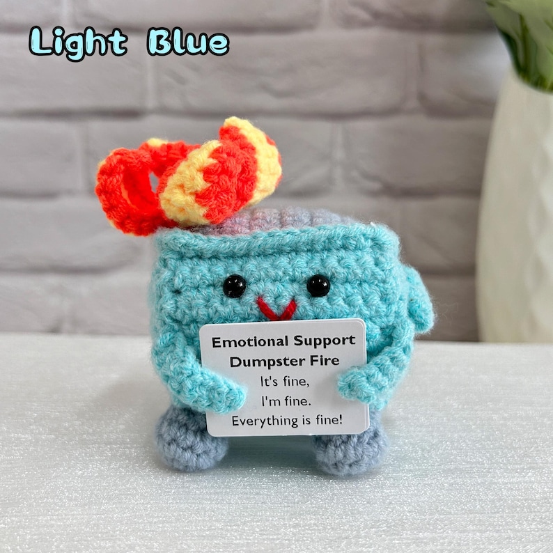 May include: A crocheted light blue dumpster fire with a red and yellow flame on top. The dumpster fire is holding a sign that says "Emotional Support Dumpster Fire It's fine, I'm fine. Everything is fine!"