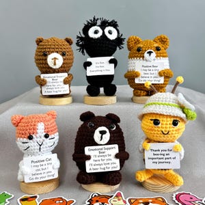May include: A selection of handmade crochet figures, including bears, a cat, and a bee, each holding a small sign with encouraging messages. The figures are in various colours, such as brown, black, orange, and yellow, and are displayed on wooden bases.