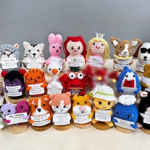 May include: A collection of handmade crochet animal figurines, including a sheep, cat, bunny, goose, crab, tiger, shark, and hamster. Each figure holds a small sign with positive messages. The figurines are displayed on wooden bases.