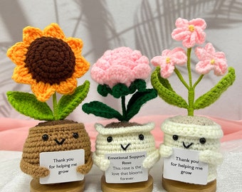 May include: Three crocheted flower pot dolls. The sunflower has orange petals and a brown center. The pink rose has green leaves. The pink flower has three blooms. Each doll holds a sign with text.