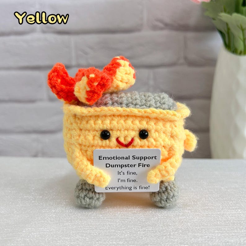 May include: A crocheted yellow dumpster fire with a red and orange flame on top. The dumpster fire has a smiling face and is holding a card that says "Emotional Support Dumpster Fire. It's fine, I'm fine. Everything is fine!"