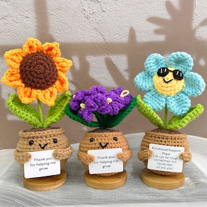 May include: Three crocheted potted plants with happy faces. The first plant is a sunflower with a brown center and orange petals. The second plant is purple with a cluster of flowers. The third plant is blue with a yellow center and sunglasses. Each plant has a sign with a message of encouragement.