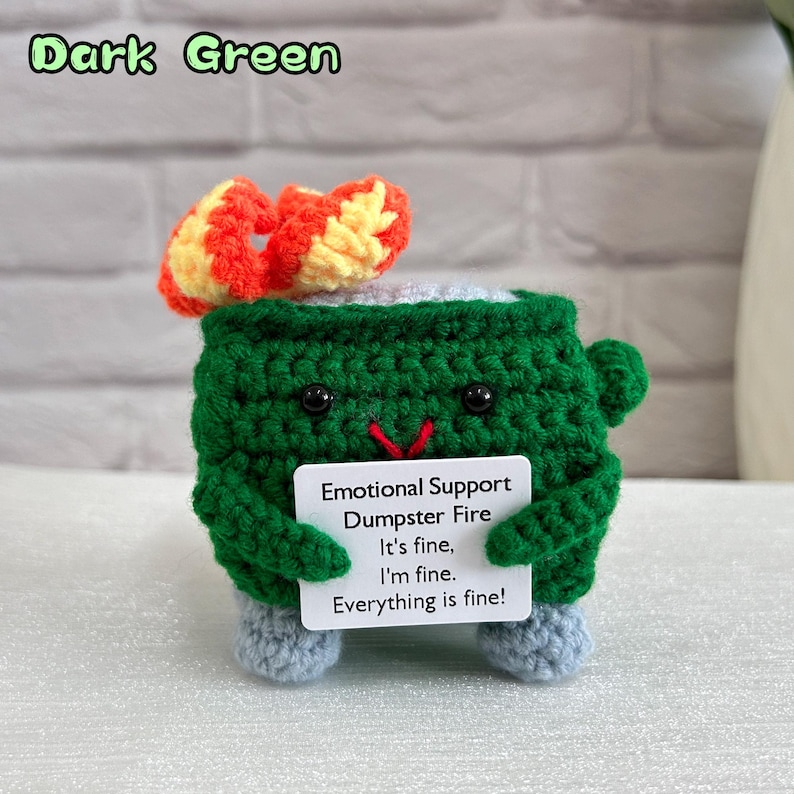 May include: A crocheted plush toy shaped like a dumpster fire with a dark green body, a red mouth, and black eyes. The toy is holding a white sign that reads "Emotional Support Dumpster Fire It's fine, I'm fine. Everything is fine!"