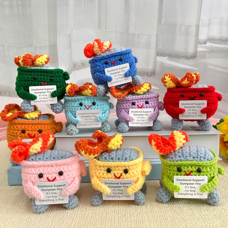 May include: Nine crocheted plush toys shaped like a dumpster fire with a smiling face and a sign that reads "Emotional Support Dumpster Fire It's fine. I'm fine. Everything is fine!" The toys are in various colors including green, blue, purple, orange, pink, yellow, and red.