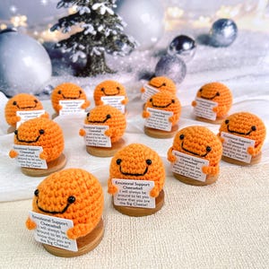 Puede incluir: "Emotional Support Cheeseball" de ganchillo naranja con una cara sonriente, sosteniendo un letrero. El letrero dice "¡Siempre estaré cerca para que sepas que eres el Big Cheese!" sobre una base de madera.