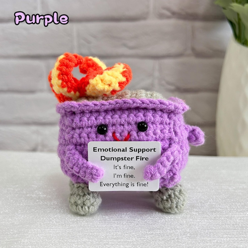 May include: Purple crocheted plush toy shaped like a dumpster fire with a red and yellow crocheted flame on top. The toy is holding a white card that says "Emotional Support Dumpster Fire It's fine, I'm fine. Everything is fine!"