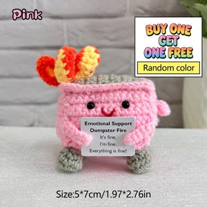 May include: A pink crocheted succulent plant with orange and yellow accents, featuring a smiling face and gray feet. It holds a sign that reads "Emotional Support Dumpster Fire." The item measures 1.97 x 2.76 inches. The image also includes the text "BUY ONE GET ONE FREE."