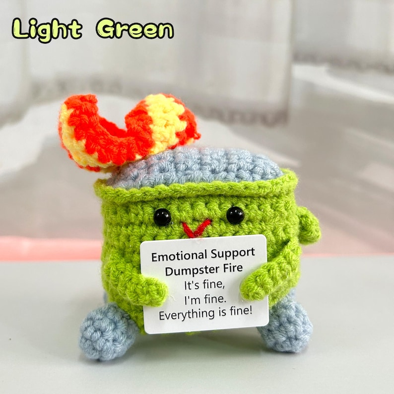 May include: A crocheted plush toy shaped like a dumpster fire with a light green body, blue lid, and orange and yellow flames. The toy is holding a white sign that reads "Emotional Support Dumpster Fire It's fine, I'm fine. Everything is fine!"