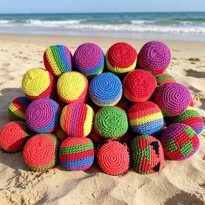 May include: A collection of colorful, crocheted hacky sacks in various colors, including red, yellow, blue, green, and purple. The hacky sacks are arranged on a sandy beach with the ocean in the background. The balls are round and appear to be handmade.