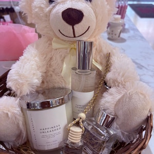 May include: A cream teddy bear in a woven basket holds a candle, perfume bottles, and a small fragrance vial. The candle has a silver rim and the text "Happiness Unleashed". The perfume bottles have silver caps and white labels.