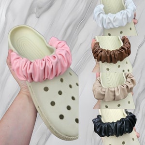 May include: Five pairs of colourful, fabric shoe charms in different colours, including white, brown, pink, beige, and black. The charms are made of a soft, stretchy fabric and are designed to fit around the back strap of a pair of clogs.