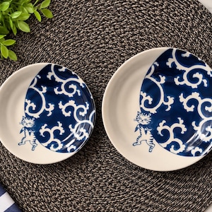 May include: Two round ceramic plates featuring a white base and a dark blue circular section decorated with white swirling patterns. Each plate displays a small, standing cat illustration in dark blue on the edge of the blue design.