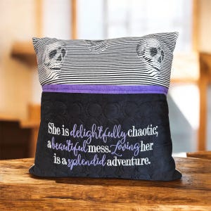 May include: Decorative pillow with a black and white striped top featuring skull imagery. The bottom portion is black with embroidered text in purple that reads "She is delightfully chaotic; a beautiful mess. Loving her is a splendid adventure."
