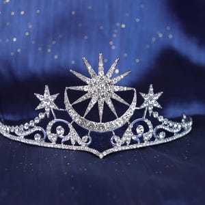 Puede incluir: Una tiara plateada con un diseño celestial que presenta un sol central, una luna creciente y estrellas. La corona está adornada con cristales brillantes y colocada sobre un fondo azul oscuro.