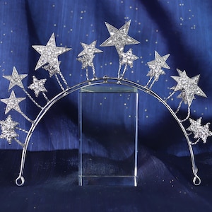 Può includere: Una tiara argentata a forma di stella con decorazioni a stella scintillanti di varie dimensioni. La tiara è ambientata su uno sfondo blu scuro con minuscoli puntini bianchi, creando un tema celeste. Le stelle sono incastonate con cristalli trasparenti.
