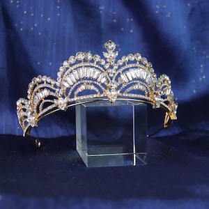 Puede incluir: Una tiara dorada adornada con cristales claros y brillantes y una gema central prominente. La tiara presenta un diseño arqueado con una serie de cristales más pequeños y piedras de talla baguette. Se muestra sobre un soporte rectangular transparente.