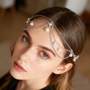 May include: A silver headpiece featuring faux pearls and small clear rhinestones. The headpiece, made of delicate chains, is worn across the forehead. The white pearls and clear rhinestones create a decorative hair accessory. The design is elegant and suitable for special occasions.