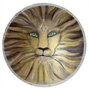 Puede incluir: Impresión artística digital enmarcada que presenta la cara de un león con melena dorada y ojos verdes penetrantes. La obra de arte circular tiene un borde decorativo y un marco dorado. Texto en la parte inferior de la imagen.