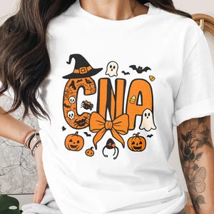 以下が含まれることがあります： ハロウィーンをテーマにしたデザインの白いTシャツ。「CNA」の文字がオレンジ色で、魔女の帽子、カボチャ、幽霊、リボンなどのハロウィーンの要素で飾られています。Tシャツはクルーネックで、袖はロールアップされています。