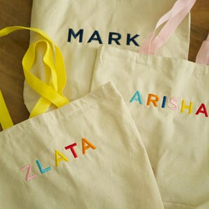May include: Three white canvas tote bags with different names embroidered on them in colorful thread. The names are "Zlata", "Arisha", and "Mark".