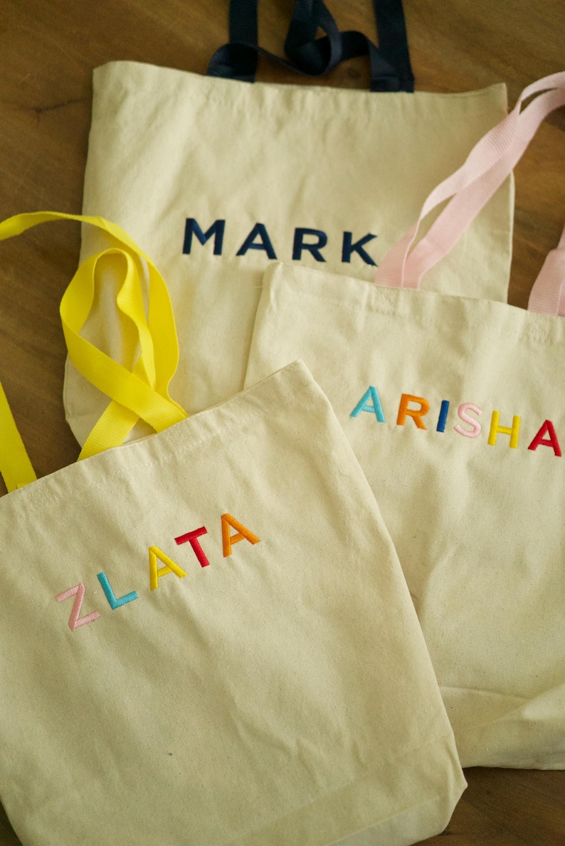 May include: Three white canvas tote bags with different names embroidered on them in colorful thread. The names are "Zlata", "Arisha", and "Mark".