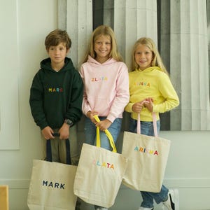 May include: Three children are holding personalized tote bags. The bags are made of natural canvas and have colorful embroidered names: "Mark", "Zlata", and "Arisha". The children are wearing matching hoodies with the same names embroidered on the front.