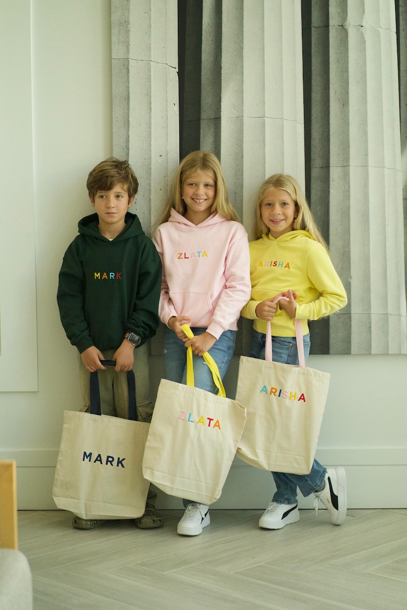 May include: Three children are holding personalized tote bags. The bags are made of natural canvas and have colorful embroidered names: "Mark", "Zlata", and "Arisha". The children are wearing matching hoodies with the same names embroidered on the front.