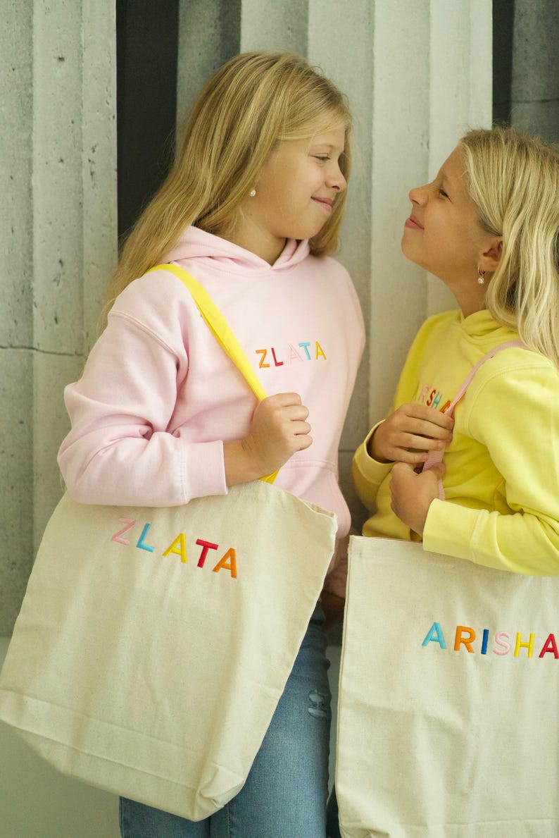 May include: Two girls are holding colorful tote bags with their names embroidered on them. The girl on the left is wearing a pink hoodie with the name "ZLATA" embroidered on it. The girl on the right is wearing a yellow hoodie with the name "ARISHA" embroidered on it.