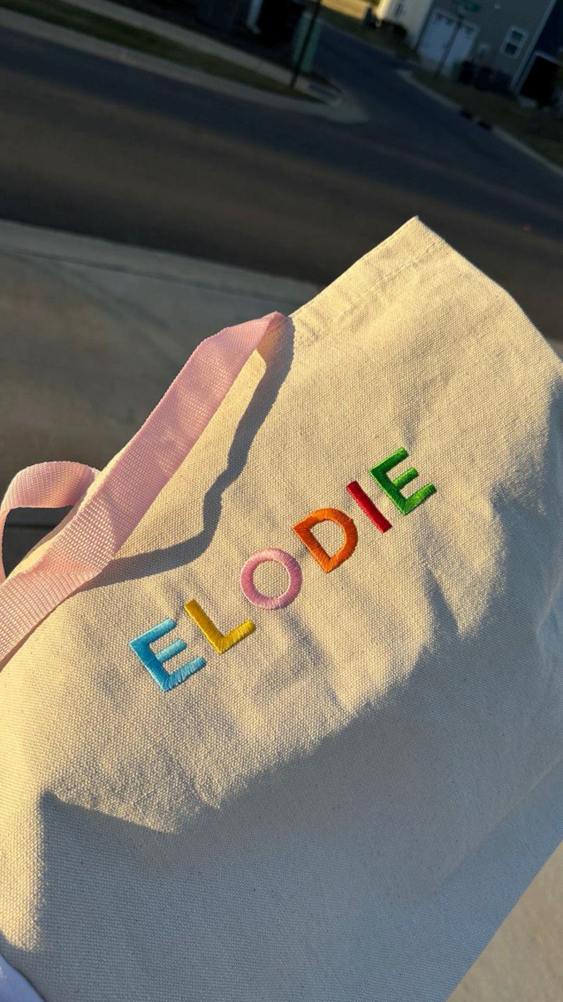 May include: A white canvas tote bag with pink straps and the name "ELODIE" embroidered in rainbow colors.