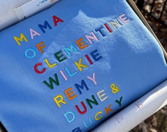 May include: A light blue sweatshirt embroidered with the words "MAMA OF CLEMENTINE WILKIE REMY DUNE & BUCKY" in various colors. The sweatshirt is inside a cardboard box with tissue paper. The text "hardest thing at the same time" is also visible.