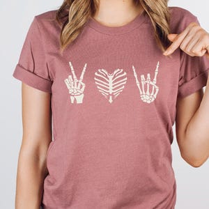 May include: Dusty rose t-shirt featuring a design with three skeletal hand gestures: a peace sign, a heart, and a rock-and-roll hand. The shirt has rolled-up sleeves and a crew neck. The design is in a cream color.