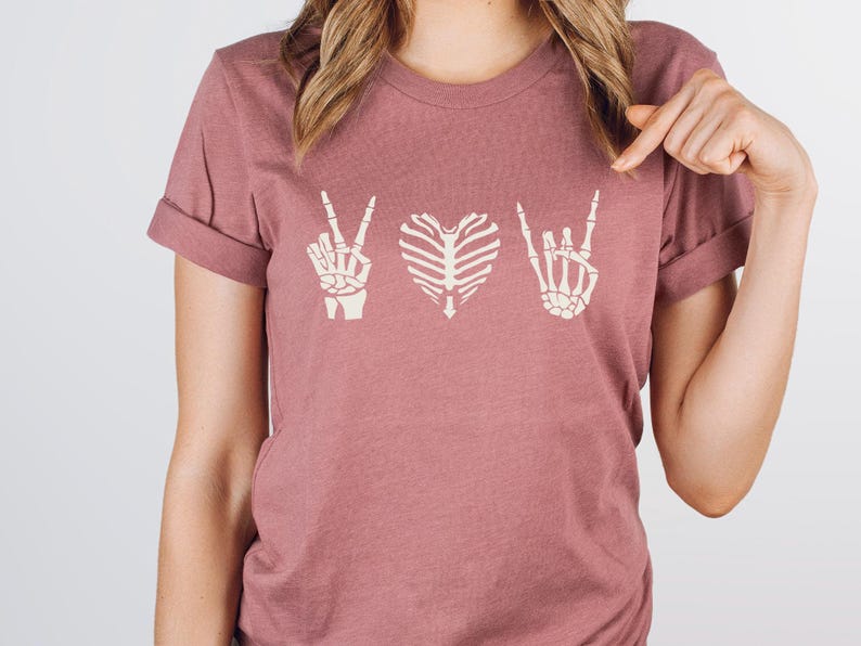 May include: Dusty rose t-shirt featuring a design with three skeletal hand gestures: a peace sign, a heart, and a rock-and-roll hand. The shirt has rolled-up sleeves and a crew neck. The design is in a cream color.