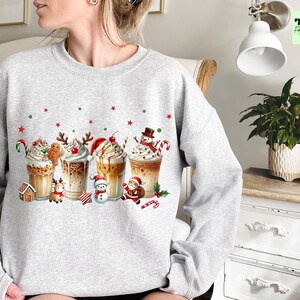 May include: A light gray sweatshirt featuring a festive holiday design. The graphic showcases several decorated coffee drinks with whipped cream, candy canes, and Christmas characters like a snowman and Santa Claus. Red and green stars and dots are scattered across the design.