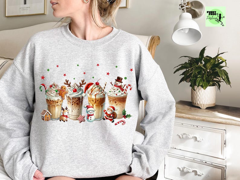May include: A light gray sweatshirt featuring a festive holiday design. The graphic showcases several decorated coffee drinks with whipped cream, candy canes, and Christmas characters like a snowman and Santa Claus. Red and green stars and dots are scattered across the design.