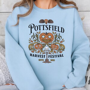 May include: A light blue crewneck sweatshirt featuring a graphic print with the text POTTSFIELD HARVEST FESTIVAL ESTD 1802. The design includes three orange jack-o'-lanterns, a small skull, autumn leaves, corn stalks, and a shovel illustration.