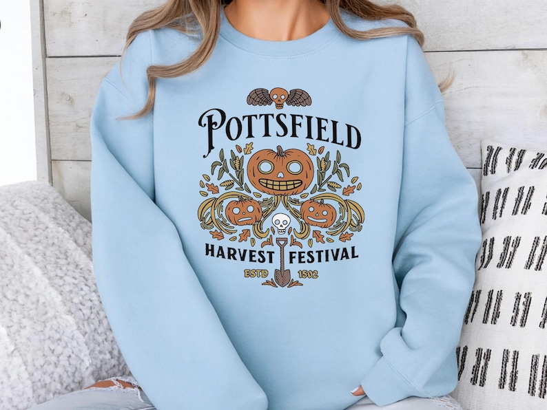 May include: A light blue crewneck sweatshirt featuring a graphic print with the text POTTSFIELD HARVEST FESTIVAL ESTD 1802. The design includes three orange jack-o'-lanterns, a small skull, autumn leaves, corn stalks, and a shovel illustration.