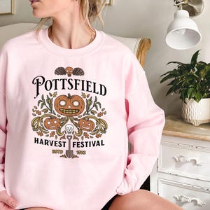 May include: A light pink crewneck sweatshirt featuring a graphic print with the text "POTTSFIELD HARVEST FESTIVAL ESTD 1902". The design includes three orange jack-o'-lanterns, autumn leaves, a small skull, and a shovel centered below.