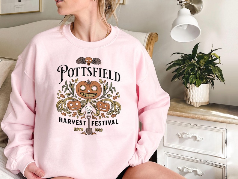 May include: A light pink crewneck sweatshirt featuring a graphic print with the text "POTTSFIELD HARVEST FESTIVAL ESTD 1902". The design includes three orange jack-o'-lanterns, autumn leaves, a small skull, and a shovel centered below.