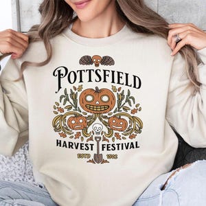 May include: A cream-colored crewneck sweatshirt featuring a graphic print with the text POTTSFIELD HARVEST FESTIVAL ESTD 1502. The design includes three carved pumpkins, a small skull, a winged skull, and autumn leaves in orange and brown.
