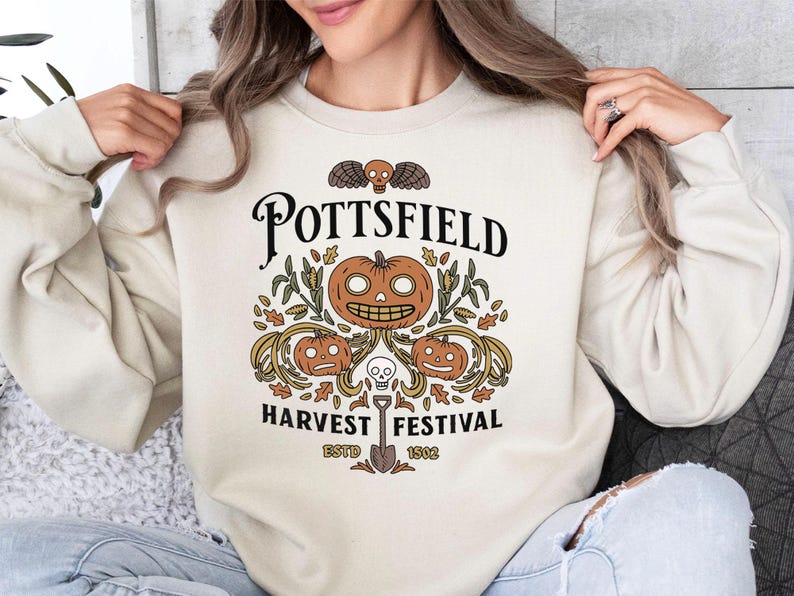 May include: A cream-colored crewneck sweatshirt featuring a graphic print with the text POTTSFIELD HARVEST FESTIVAL ESTD 1502. The design includes three carved pumpkins, a small skull, a winged skull, and autumn leaves in orange and brown.