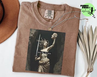May include: A brown t-shirt with a graphic of a statue holding a sword and a severed head, with the text "Girls Will Be Girls." A silver necklace with a pendant is draped across the shirt. The shirt has a Comfort Colors tag.