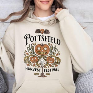 May include: A beige hooded sweatshirt featuring a graphic print with the text POTTSFIELD HARVEST FESTIVAL ESTD 1502. The design includes three orange jack-o'-lanterns, a small skull with wings, autumn leaves, and a shovel centered below.
