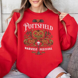 May include: A bright red long-sleeve sweatshirt featuring a graphic print with the text POTTSFIELD HARVEST FESTIVAL ESTD 1502. The design includes three orange jack-o'-lanterns, green foliage, a small skull, and a shovel in black and white ink.