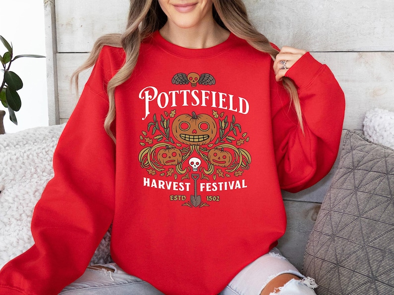 May include: A bright red long-sleeve sweatshirt featuring a graphic print with the text POTTSFIELD HARVEST FESTIVAL ESTD 1502. The design includes three orange jack-o'-lanterns, green foliage, a small skull, and a shovel in black and white ink.